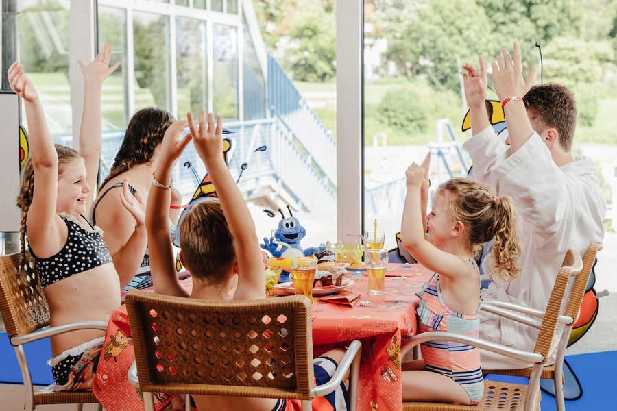 Familie sitzt im Restaurant eines Schwimmbads und wirft die Arme vor Freude nach oben.