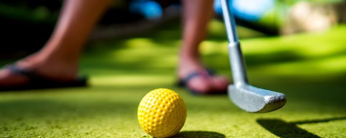 Grüner Korkboden, auf dem ein gelber Golfball liegt. Daneben ist ein Teil eines Minigolfschlägers zu sehen, der bereit ist, den Golfball einzulochen.