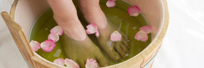 Zwei nackte Füße im Holzeimer, der mit Wasser und rosafarbenen Rosenblättern befüllt ist