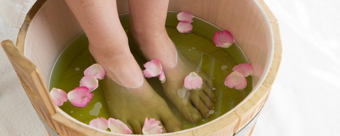 2 nackte Füße im Holzeimer, der mit Wasser und rosafarbenen Rosenblättern befüllt ist