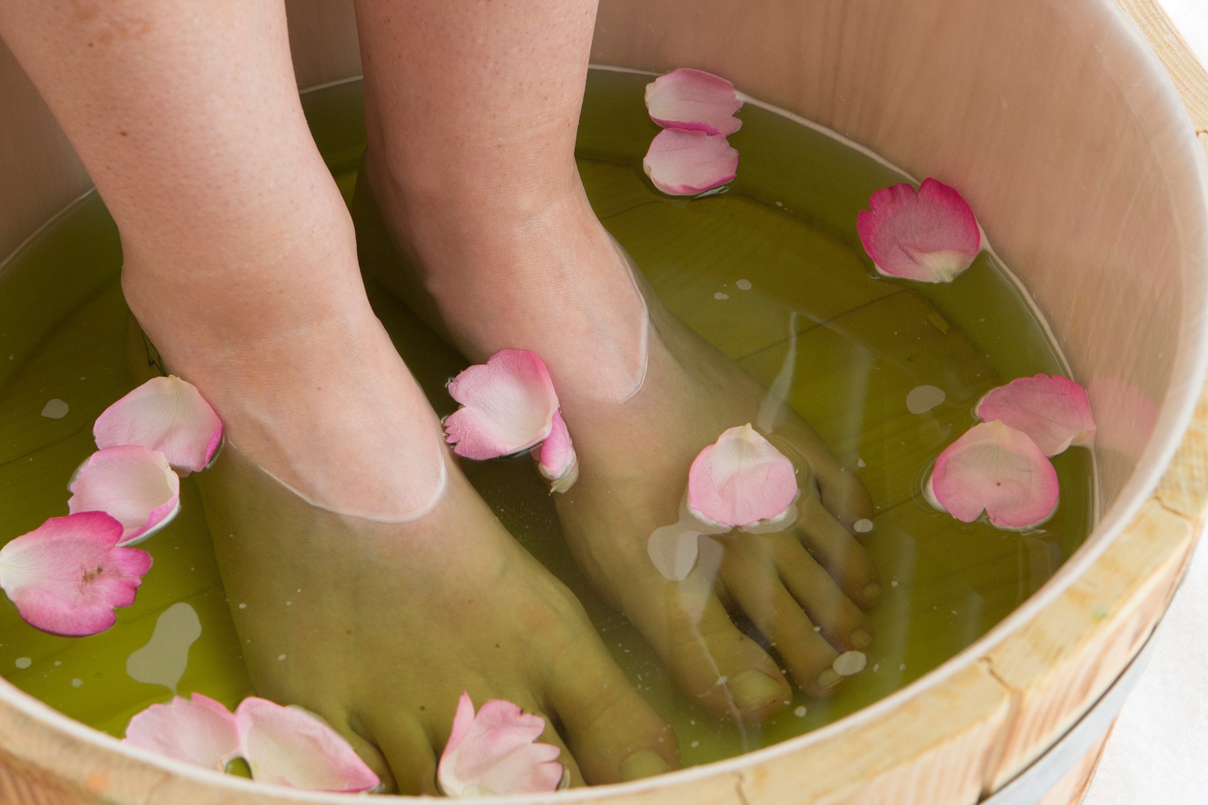 Fußpflege in der Copa Oase Zwei nackte Füße im Holzeimer, der mit Wasser und rosafarbenen Rosenblättern befüllt ist