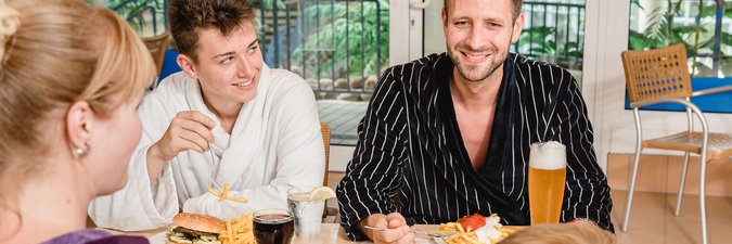 Zwei Männer und eine Frau sitzen im Restaurantbereich. Sie trinken Bier und Cola und essen einen Hamburger beziehungsweise eine Pommes rot-weiss.