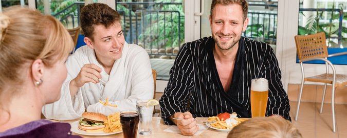 Zwei Männer und eine Frau sitzen im Restaurantbereich. Sie trinken Bier und Cola und essen einen Hamburger beziehungsweise eine Pommes rot-weiss.