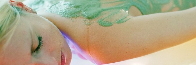 blonde Frau liegt auf dem Bauch auf einer Liege und hat die Augen geschlossen. Auf ihrem Rücken ist eine grüner Schlamm verteilt. Sie hat die Augen geschlossen