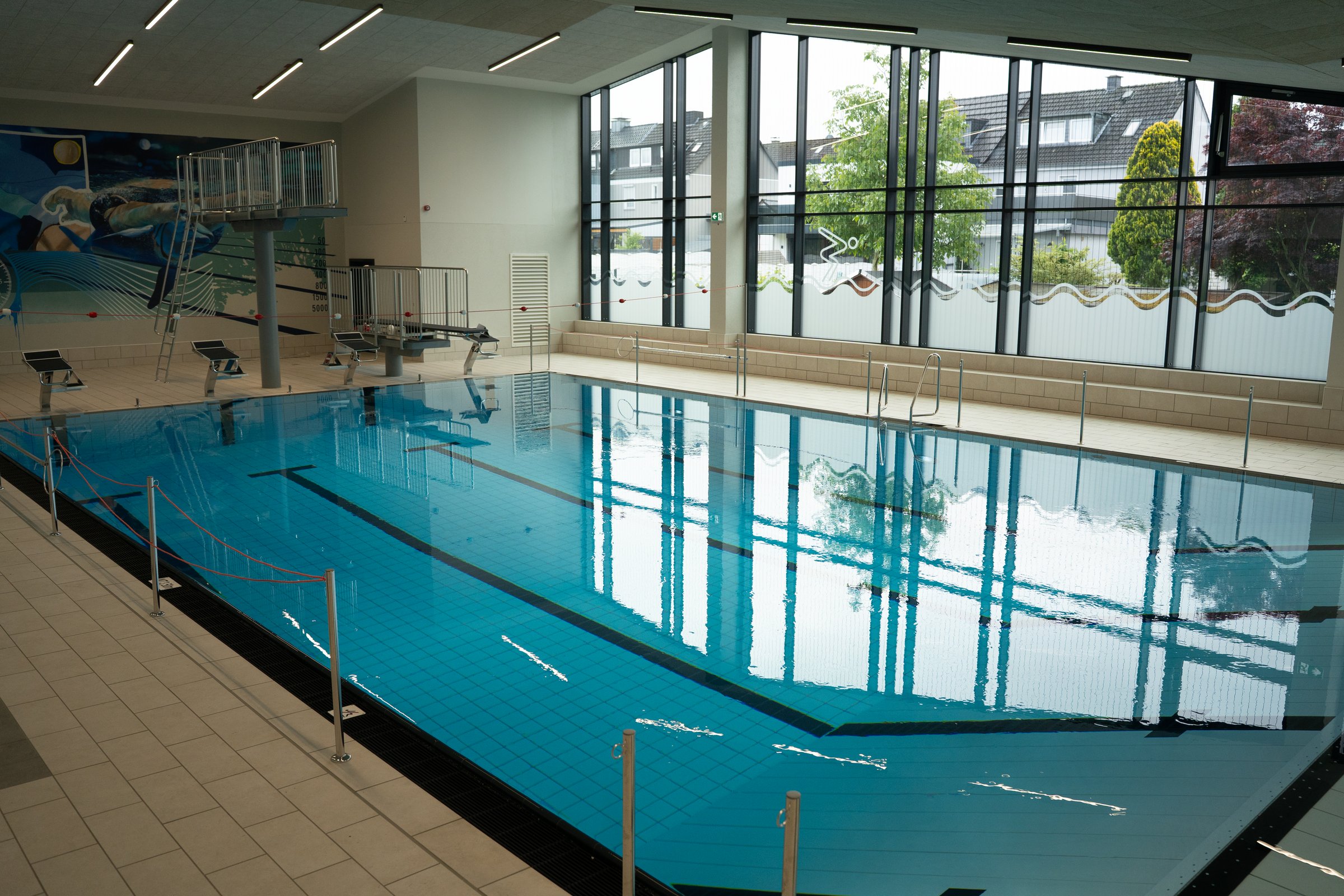 Weitwinkelaufnahme über das Schwimmerbecken mit Sprungturm, im Hintergrund Glasfassade. 
