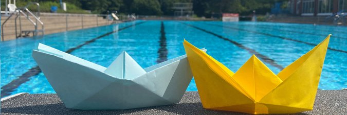 Zwei Origami-gefaltete Papierboote stehen auf dem Beckenrand eines Schwimmbeckens im Freibad.