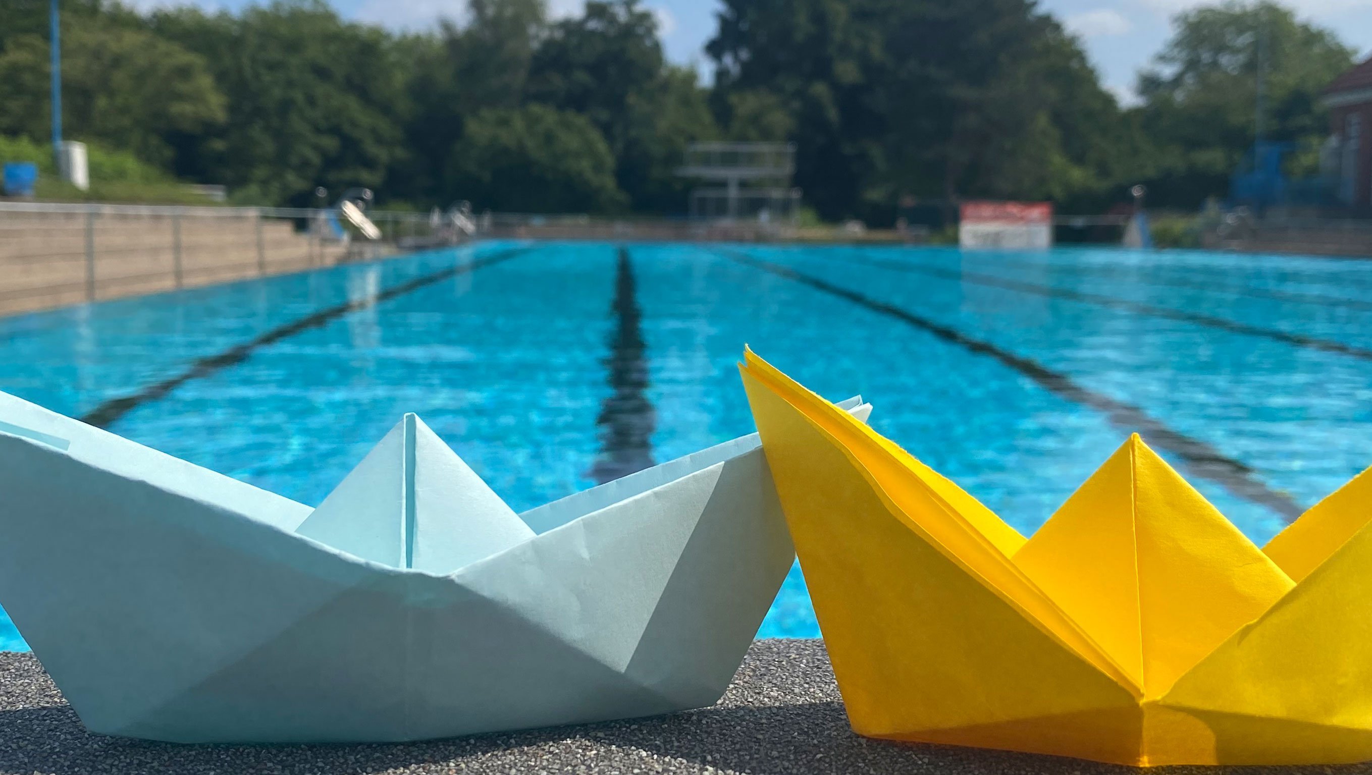 Zwei Origami-gefaltete Papierboote stehen auf dem Beckenrand eines Schwimmbeckens im Freibad.