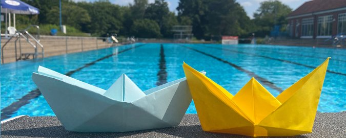 Zwei Origami-gefaltete Papierboote stehen auf dem Beckenrand eines Schwimmbeckens im Freibad.