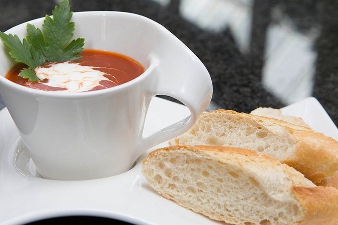 Ein geschwungener weißer Teller, auf dem ein helles Stück Brot liegt. Daneben befindet sich eine passende weiße Tasse mit einer roten Tomatensuppe und einem Klecks weißer Sahne mit einem grünen Minzzweig