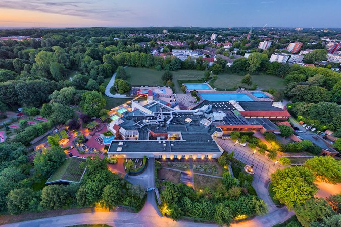 Ein Schwimmbad von oben, zwischen Bäumen am Stadtrand lokalisiert. Im Hintergrund ein in rot und blau geteilter Himmel.