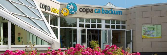 Eingang Copa ca backum Eingangsbereich von außen, weiße Konstruktion mit Glas verkleidet, im Vordergrund befinden sich rote Blumen