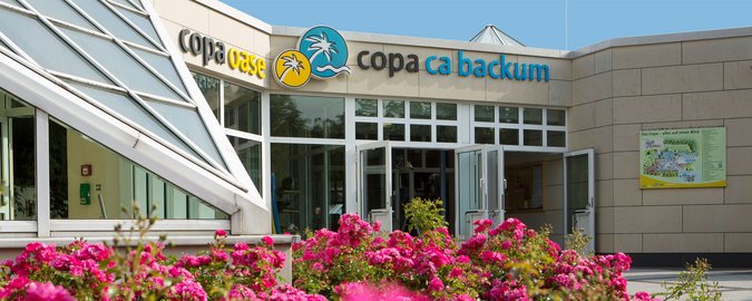 Eingang Copa ca backum Eingangsbereich von außen, weiße Konstruktion mit Glas verkleidet, im Vordergrund befinden sich rote Blumen