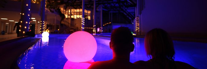 Ein Paar sitzt am Rand eines großen, blau beleuchteten Schwimmbeckens, auf der Wasseroberfläche treiben bunte Leuchten. Es ist weitgehend dunkel, nur im Hintergrund sind wenige Lichter und Kerzen zu sehen.