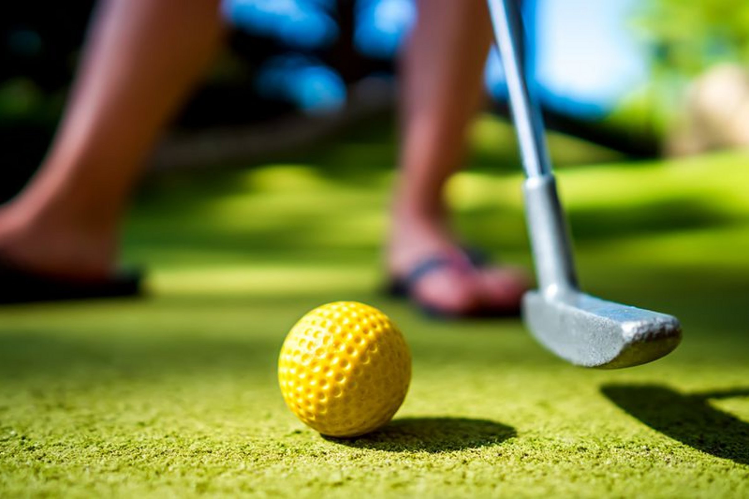 Minigolf am Copa Ca Backum Grüner Korkboden, auf dem ein gelber Golfball liegt. Daneben ist ein Teil eines Minigolfschlägers zu sehen, der bereit ist, den Golfball einzulochen.