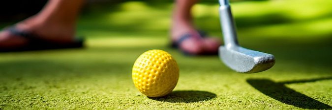 Grüner Korkboden, auf dem ein gelber Golfball liegt. Daneben ist ein Teil eines Minigolfschlägers zu sehen, der bereit ist, den Golfball einzulochen.