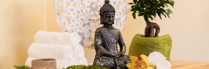heller Holztisch auf dem eine kleine schwarze Buddhafigur und ein kleines Bonsaibäumchen in einem grünen Topf steht. Daneben befindet sich ein Stapel weißer Handtücher und eine Messingschale