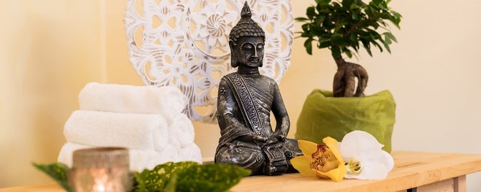 heller Holztisch auf dem eine kleine schwarze Buddhafigur und ein kleines Bonsaibäumchen in einem grünen Topf steht. Daneben befindet sich ein Stapel weißer Handtücher und eine Messingschale
