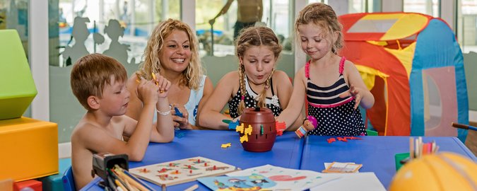 Eine blonde Frau und drei Kinder spielen an einem blauen Tisch ein Gesellschaftsspiel.