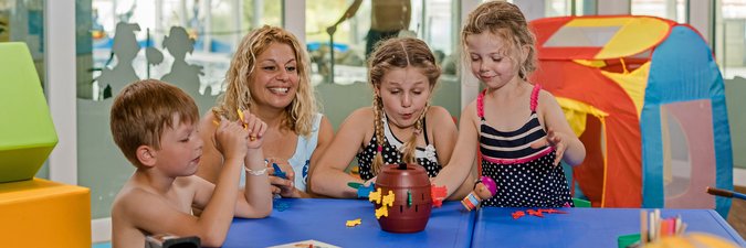 Eine blonde Frau und drei Kinder spielen an einem blauen Tisch ein Gesellschaftsspiel.