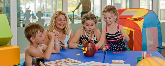 Eine blonde Frau und drei Kinder spielen an einem blauen Tisch ein Gesellschaftsspiel.