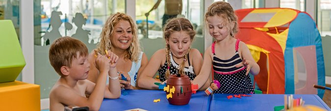 Eine blonde Frau und drei Kinder spielen an einem blauen Tisch ein Gesellschaftsspiel.