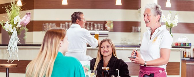 Eine Kellnerin mit weißer Bluse und roter Schürze bedient mit guter Laune zwei Frauen, die an einem Tisch im Restaurant sitzen und Cola trinken.