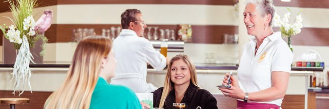 Eine Kellnerin mit weißer Bluse und roter Schürze bedient mit guter Laune zwei Frauen, die an einem Tisch im Restaurant sitzen und Cola trinken.