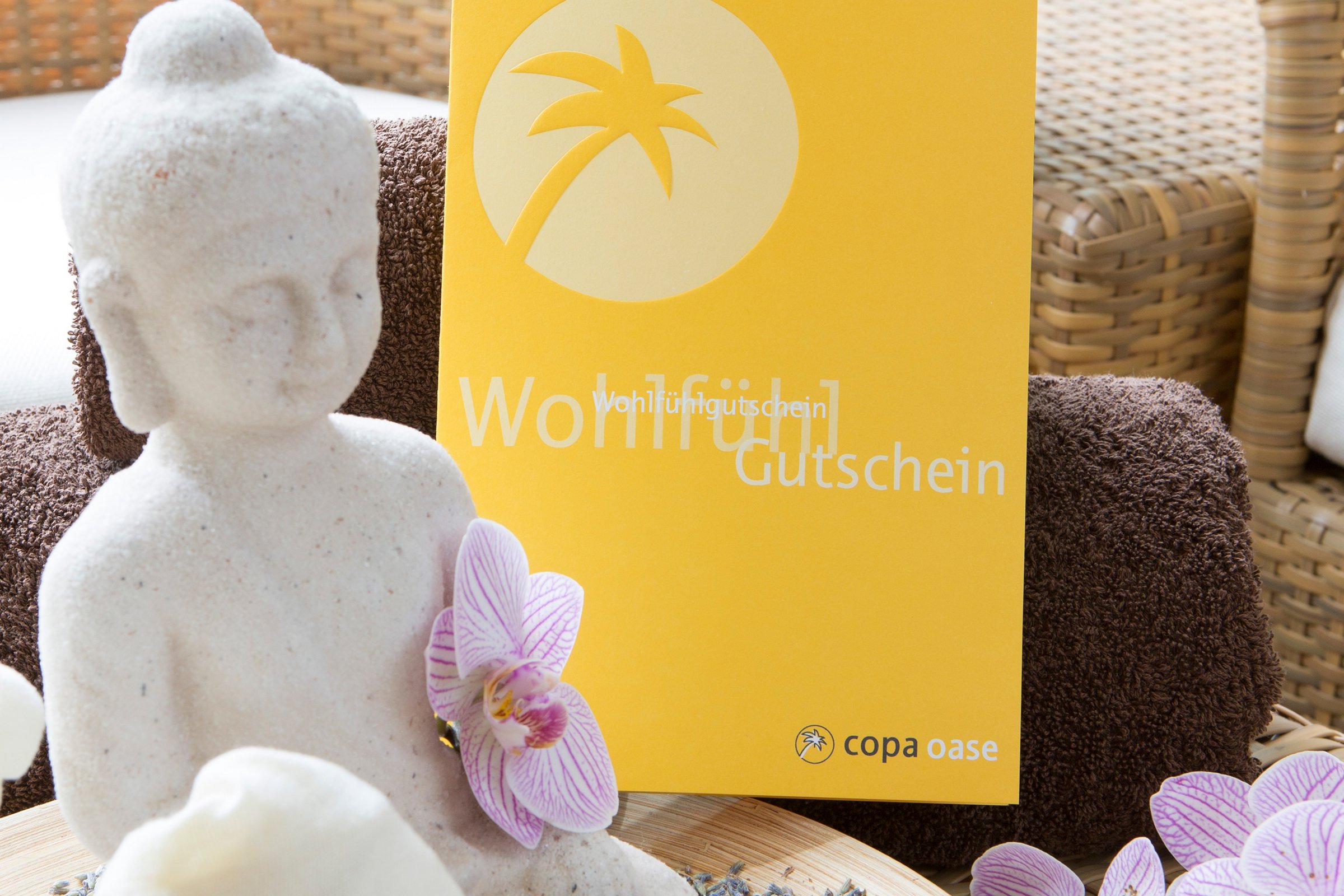 Onlineshop des Copa Ca Backum und der Copa Oase Weiße, kleine Buddhafigur sitzt auf Rattan-Tisch vor einem gelben Infoprospekt.