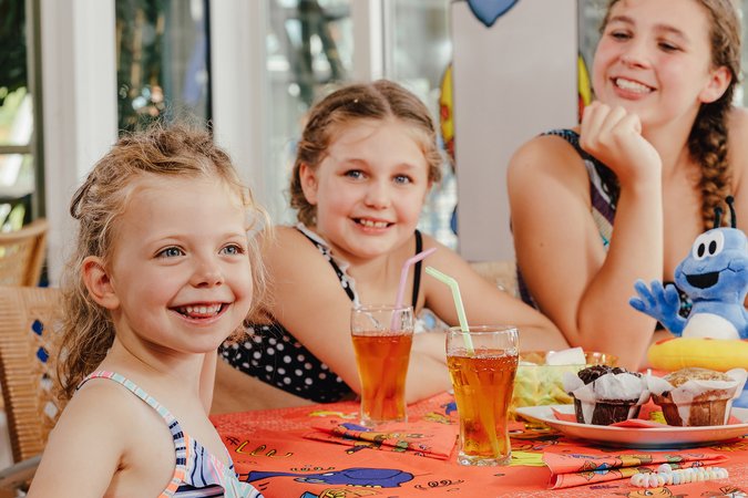 Drei junge Mädchen sitzen im Badeanzug am Tisch eines Bistros, trinken Eistee und haben zwei Muffins vor sich stehen.