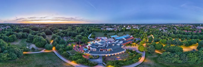 Panorama-Blick des Copa Ca Cackum aus der Vogelperspektive. Panorama-Blick auf ein Schwimmbad, ansässig mitten zwischen Wäldern und Wiesen, im Hintergund erkennt man eine Stadt.