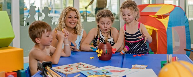 Frau sitzt mit drei Kindern am Spieletisch und sie spielen verschiedene Spiele.