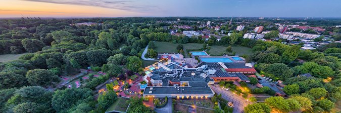 Weitwinkel-Blick auf ein Schwimmbad, ansässig mitten zwischen Wäldern und Wiesen, im Hintergund erkennt man eine Stadt zur Abenddämmerung.